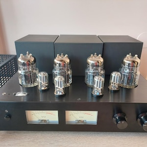 Op de afbeelding: Een zwarte buizenversterker met vier grote en vier kleine glazen vacuümbuizen op de bovenzijde. Het voorpaneel bevat twee analoge VU-meters met beige wijzerplaten, twee zwarte bedieningsknoppen en een kleine aan-uitknop aan de linkerzijde.