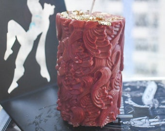 May include: A cylindrical pillar candle in a deep red hue, featuring an intricate, raised relief pattern of swirling organic shapes across its surface. The top of the candle is adorned with scattered gold leaf flakes and a single centered wick.