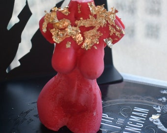 May include: A red torso-shaped candle decorated with gold leaf flakes across the upper chest and shoulder area. The wax sculpture features a smooth, matte finish and is positioned in front of a black object with the text TIP FOR THE HANGMAN.