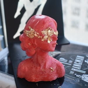 Puede incluir: Una vela de cera roja con forma de busto humano y acabado liso. La figura presenta una venda hecha de copos de pan de oro aplicados sobre los ojos y la frente. La escultura representa a una persona con un peinado estilizado y expresión neutra.