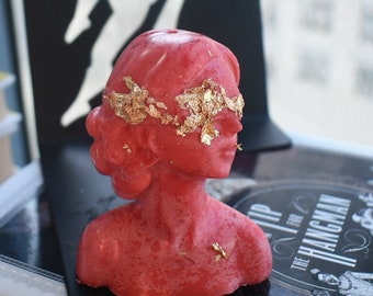 May include: A red wax candle shaped as a human bust with a smooth finish. The figure features a blindfold made of applied gold leaf flakes across the eyes and forehead. The sculpture depicts a person with a stylized hairstyle and a neutral expression.
