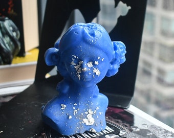 May include: A bright blue wax bust of a person with stylized hair, featuring scattered silver leaf flakes applied to the face and chest. The sculpture has a smooth, molded surface and is shaped as a head and shoulders portrait with detailed hair.