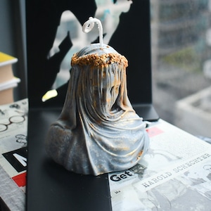Puede incluir: Una vela con forma de busto gris que representa una figura envuelta en tela con una corona pintada en oro sobre la cabeza. La vela presenta una mecha blanca que sobresale de la parte superior y detalles dorados que resaltan los pliegues.