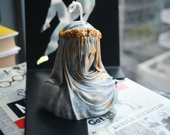 May include: A gray bust candle shaped like a figure draped in fabric with a gold-painted wreath on the head. The candle features a white wick protruding from the top and gold accents highlighting the folds of the fabric and the crown detail.