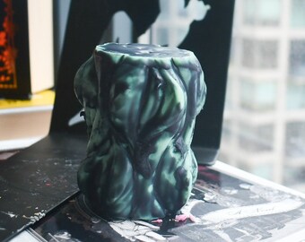 May include: A cylindrical pillar candle featuring a marbled pattern of deep forest green and black wax. The surface has a textured, organic appearance with vertical ridges and drips, creating a layered effect across the entire exterior of the candle.