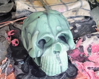 May include: A molded skull object featuring a pale mint green base color with dark gray or black marbled streaks running down from the top and pooling around the base. The skull has deep, dark eye sockets and a defined nasal cavity with a drip effect.