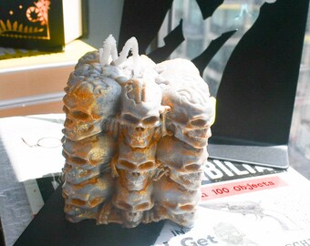 May include: A square-shaped pillar candle featuring a stacked design of multiple human skulls. The candle is colored in a muted grey with metallic gold accents highlighting the skull details. A single white wick protrudes from the top center.
