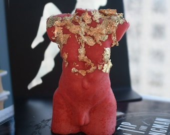 May include: A red torso-shaped candle featuring an applied gold leaf finish across the upper chest and shoulder area. The sculpture depicts a human torso with a textured surface, standing upright as a decorative object with no visible wick.