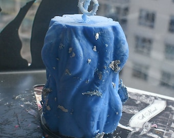 May include: A cylindrical blue candle with a textured, wavy surface and embedded silver leaf flakes. The candle features a single white wick at the top center. The wax has a matte finish and irregular vertical ridges throughout its exterior structure.