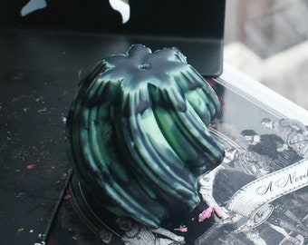 May include: A round, swirled candle featuring a blend of dark forest green and black wax. The top surface has a star-shaped indentation with a central wick hole. The wax has a glossy finish with deep, flowing vertical ridges throughout the body.