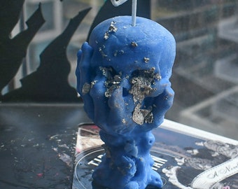 May include: A blue wax candle shaped like a human skull held by two hands. The candle features a white wick at the top and is decorated with scattered silver-colored metallic flakes embedded into the surface, particularly around the eye sockets.