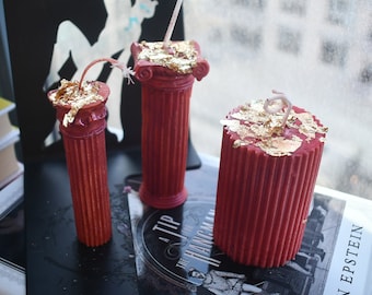 May include: Three red wax candles with gold leaf accents on top. Two candles are shaped like fluted columns with capitals, and one is a wider cylindrical pillar candle. Each candle features a single cotton wick protruding from the center top.