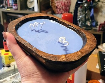 May include: An oval-shaped candle with light blue wax poured into a dark brown, rustic wooden dough bowl. The candle features two white cotton wicks positioned in the center of the smooth, solid wax surface, contained within the natural wood grain.