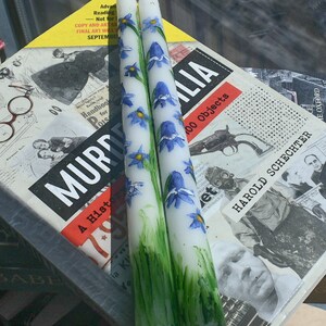 Puede incluir: Dos velas c&oacute;nicas blancas con flores azules en forma de campana pintadas a mano y hierba verde en la base. Las velas tienen una mecha blanca en la parte superior y est&aacute;n colocadas verticalmente, mostrando un dise&ntilde;o bot&aacute;nico sobre la cera blanca.