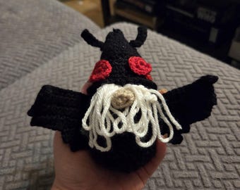 May include: A handcrafted crochet bat figure featuring a black body and wings, two prominent red circular eyes, and a long, white yarn beard. The creature has small black ears at the top and a small tan nose visible beneath the white yarn beard.