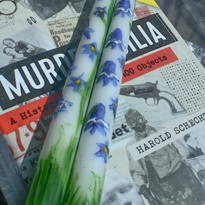 Puede incluir: Dos velas c&oacute;nicas blancas con flores azules en forma de campana pintadas a mano, con centros amarillos y tallos verdes. La base de cada vela est&aacute; decorada con pinceladas verdes verticales que representan hierba y una mecha blanca.