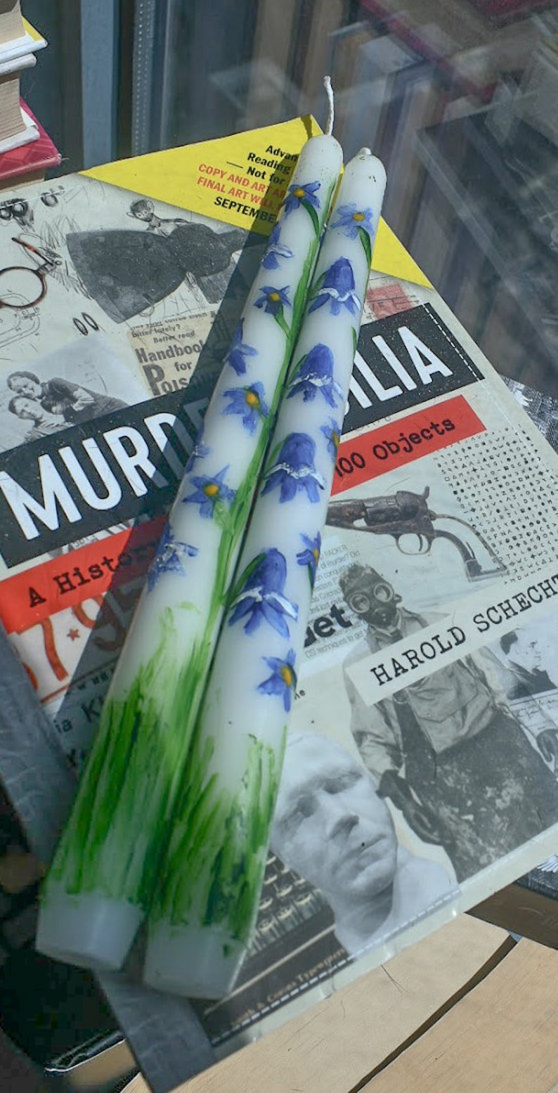 Puede incluir: Dos velas c&oacute;nicas blancas con flores azules en forma de campana pintadas a mano, con centros amarillos y tallos verdes. La base de cada vela est&aacute; decorada con pinceladas verdes verticales que representan hierba y una mecha blanca.