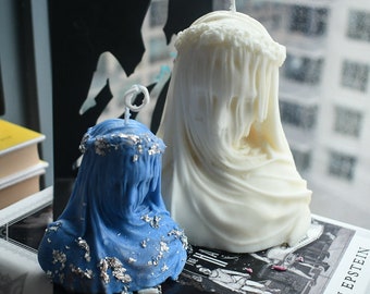 May include: Two sculptural candles shaped like draped busts. The smaller candle is blue with silver leaf accents, while the larger candle is solid white. Both feature a circular wreath detail around the head and a single vertical wick at the top.