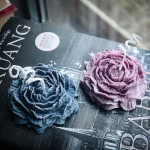 Puede incluir: Dos velas con forma de flor, una de color azul oscuro y otra de color morado claro, cada una con una mecha blanca en espiral. Las velas descansan sobre la portada de un libro negro con texto blanco que incluye la palabra KUANG y una pegatina circular.