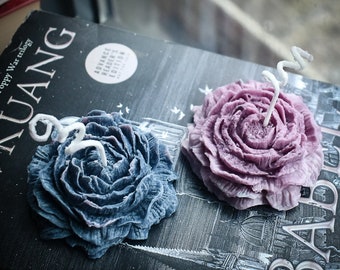 May include: Two flower-shaped candles, one in dark blue and one in light purple, each featuring a spiraled white wick. The candles are resting on a black book cover with white text that includes the word KUANG and a circular sticker.