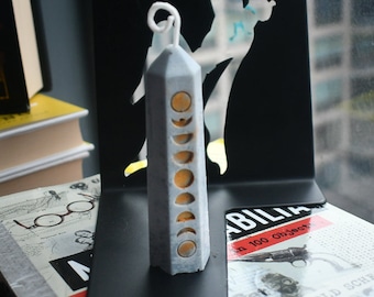 May include: A light gray, hexagonal pillar candle featuring a vertical sequence of golden moon phase icons along one side. The candle has a white, curled wick at the top and stands upright on a flat surface, displaying a smooth, matte finish.