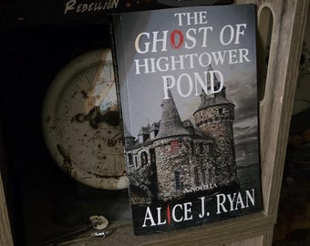 May include: A paperback book titled The Ghost of Hightower Pond by Alice J. Ryan, featuring a cover illustration of a stone castle with turrets under a dark sky, positioned on a shelf alongside other books and a vintage round scale.