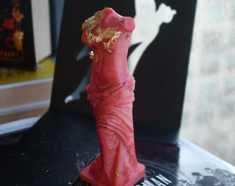 May include: A red wax candle shaped like a classical torso sculpture, featuring draped fabric details and irregular gold leaf flakes applied to the upper chest and shoulder area. The candle stands on a square base and has a smooth, matte finish.