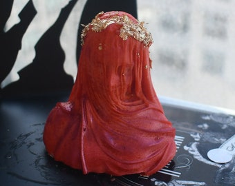 May include: A red bust sculpture depicting a figure draped in fabric, featuring a crown-like arrangement of gold leaf flakes on the head. The sculpture has a smooth, matte texture and shows intricate folds in the material covering the face and shoulders.