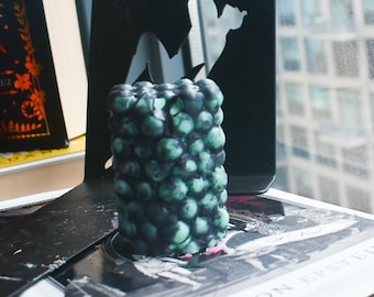 May include: A cylindrical pillar candle with a textured surface composed of numerous small, rounded spheres. The candle features a marbled color pattern blending deep black and muted seafoam green tones throughout its entire exterior structure.