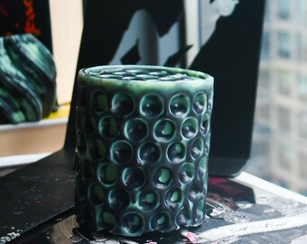 May include: A cylindrical candle featuring a dark green and black honeycomb-like exterior texture. The wax surface displays a repeating pattern of deep, circular indentations that create a porous, three-dimensional appearance across the entire body.