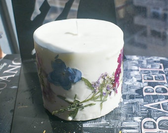 May include: A cylindrical white wax candle featuring embedded dried flowers in shades of blue, purple, and pink. The candle has a single centered wick on the top surface and is decorated with pressed botanical elements around its exterior side.