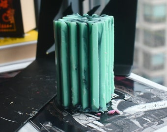 May include: A cylindrical pillar candle featuring a textured, fluted exterior surface in a pale mint green color with dark charcoal gray marbling streaks running vertically throughout the wax structure, creating a distinct ribbed appearance.