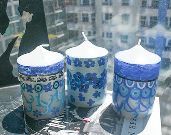 May include: Three cylindrical white wax candles featuring hand-painted blue designs. The left candle has a blue border and floral motifs. The center candle displays small blue flowers. The right candle shows a blue scalloped pattern with lines.