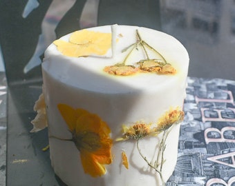 May include: A cylindrical white wax candle featuring embedded dried yellow and orange flowers on its top and side surfaces. The candle has a single centered wick and is positioned against a dark background with partial text visible on the right side.