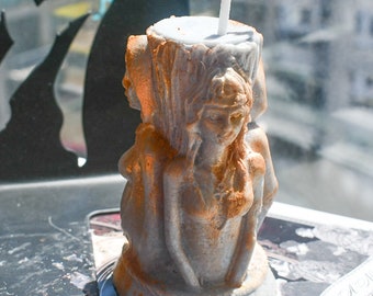May include: A cylindrical gray wax candle featuring three sculpted female figures in relief around the exterior. The figures are accented with metallic gold paint. A single white cotton wick protrudes from the flat top of the candle.