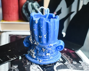 May include: A bright blue pillar candle shaped like a classical architectural column with a fluted top and scroll-like volutes at the base. The wax surface is decorated with scattered silver leaf flakes and features a single white cotton wick at the center.