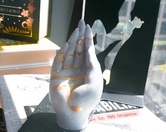 May include: A light gray hand-shaped candle with a single white wick extending from the top. The palm and fingers are decorated with metallic gold paint accents in horizontal lines and strokes across the surface of the wax sculpture.