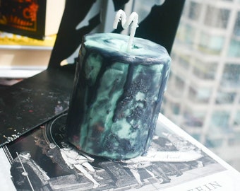 May include: A cylindrical pillar candle featuring a marbled blend of dark charcoal black and pale mint green wax. The surface has a textured, dripped appearance with dark wax running down the sides. A single white wick protrudes from the top center.