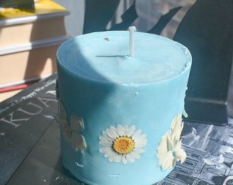 May include: A cylindrical light blue pillar candle featuring embedded pressed white daisy flowers with yellow centers on the exterior surface. The candle has a single centered white wick protruding from the top flat surface of the wax.