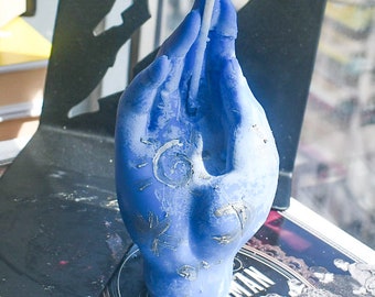 May include: A blue wax candle shaped like a hand with fingers pointing upward. The candle features a white wick extending from the center and silver-colored etched designs on the palm, including a sun, a spiral, and other celestial-like symbols.