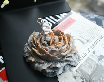 May include: A gray flower-shaped candle with gold-toned accents on the petal edges. The candle features a white, coiled wick extending from the center. The textured, layered petals mimic the appearance of a blooming flower in a circular form.
