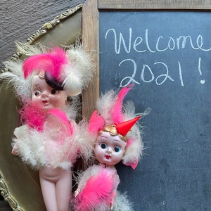 May include: Two vintage dolls dressed in pink and white feathered costumes. The dolls are standing in front of a chalkboard with the words "Welcome 2021!" written in white chalk.