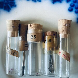 May include: Five glass vials with cork stoppers and labels. The labels have numbers on them, including 25, 1315, 8, and 20/120. The vials contain small metal objects.