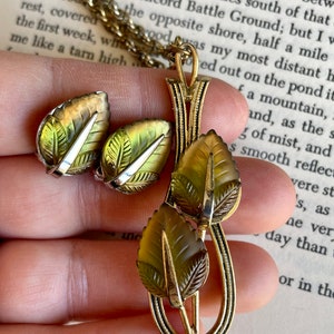 K&ouml;nnte beinhalten: Ein Vintage-Schmuckset mit einem goldfarbenen Kettenanh&auml;nger und passenden Clip-Ohrringen. Der Anh&auml;nger und die Ohrringe sind mit Blattmotiven in Gr&uuml;n- und Braunt&ouml;nen mit silbernen Akzenten gestaltet. Das Set wird an einer Kette pr&auml;sentiert.