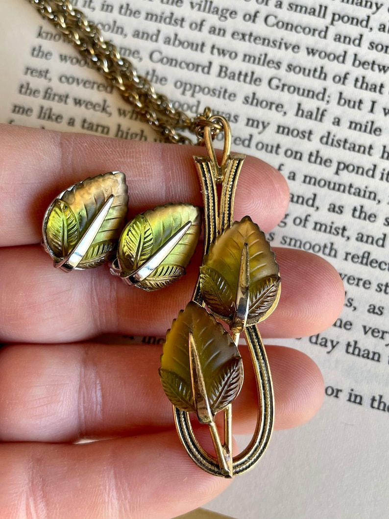 K&ouml;nnte beinhalten: Ein Vintage-Schmuckset mit einem goldfarbenen Kettenanh&auml;nger und passenden Clip-Ohrringen. Der Anh&auml;nger und die Ohrringe sind mit Blattmotiven in Gr&uuml;n- und Braunt&ouml;nen mit silbernen Akzenten gestaltet. Das Set wird an einer Kette pr&auml;sentiert.