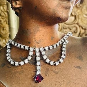 May include: A silver-toned necklace with clear, round gemstones and a dangling, square, red gemstone. The necklace is displayed on a bronze-colored statue, showcasing its elegant design and sparkling details.