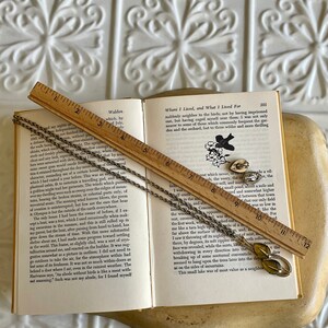 K&ouml;nnte beinhalten: Ein Vintage-Schmuckset mit Halskette und Ohrringen mit Blattdesign, das auf einem offenen Buch mit einem Holzlineal liegt. Der Schmuck hat eine silberfarbene Oberfl&auml;che mit gr&uuml;nen und gelben Akzenten. Das Buch zeigt gedruckten Text.