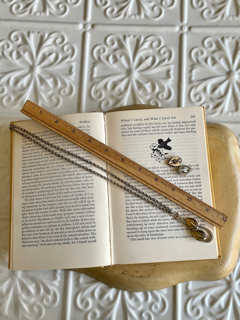 K&ouml;nnte beinhalten: Ein Vintage-Schmuckset mit Halskette und Ohrringen mit Blattdesign, das auf einem offenen Buch mit einem Holzlineal liegt. Der Schmuck hat eine silberfarbene Oberfl&auml;che mit gr&uuml;nen und gelben Akzenten. Das Buch zeigt gedruckten Text.