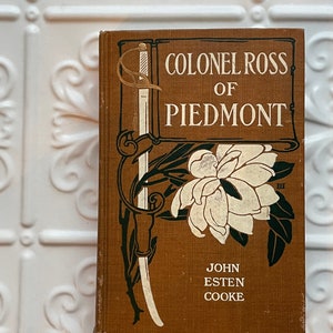 May include: A brown book with a gold and black illustration of a sword and a flower. The title is "Colonel Ross of Piedmont" by John Esten Cooke.