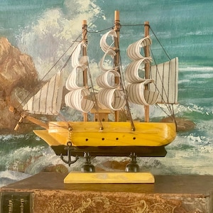 May include: A model ship featuring a bright yellow hull with a black base, three wooden masts, and white fabric sails with thin brown stripes. A small black metal anchor hangs from the bow, and the vessel is mounted on a rectangular wooden stand.
